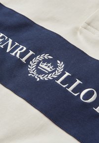 Tessuto di cotone con una combinazione di colori crema e navy. Presenta il testo ricamato "HENRI LLOYD" e un design a corona d'alloro.