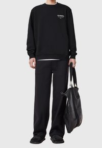 Persona con sudadera y pantalones negros, sosteniendo un gran bolso de cuero negro con correas dobles para el hombro.