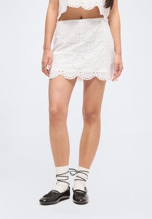 Naine, kes kannab valget õõnespitsiga seelikut ja lühikest topi, mida on kombineeritud valgete sokkide ja mustade paeltega baleriinadega.