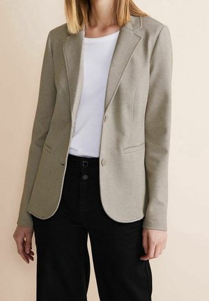 Mujer con blazer beige sobre camisa blanca y pantalones negros, de pie frente a un fondo beige liso.