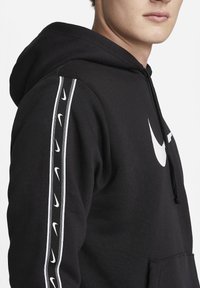 Junge Person trägt einen schwarzen Nike-Hoodie mit weißen Nike-Logos auf der Brust und am Ärmel, stehend vor weißem Hintergrund.