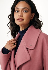 Cappotto in misto lana rosa con grandi revers, indossato sopra un dolcevita blu navy. Mano tocca il colletto, anello d'oro sulla mano sinistra, tessuto strutturato.