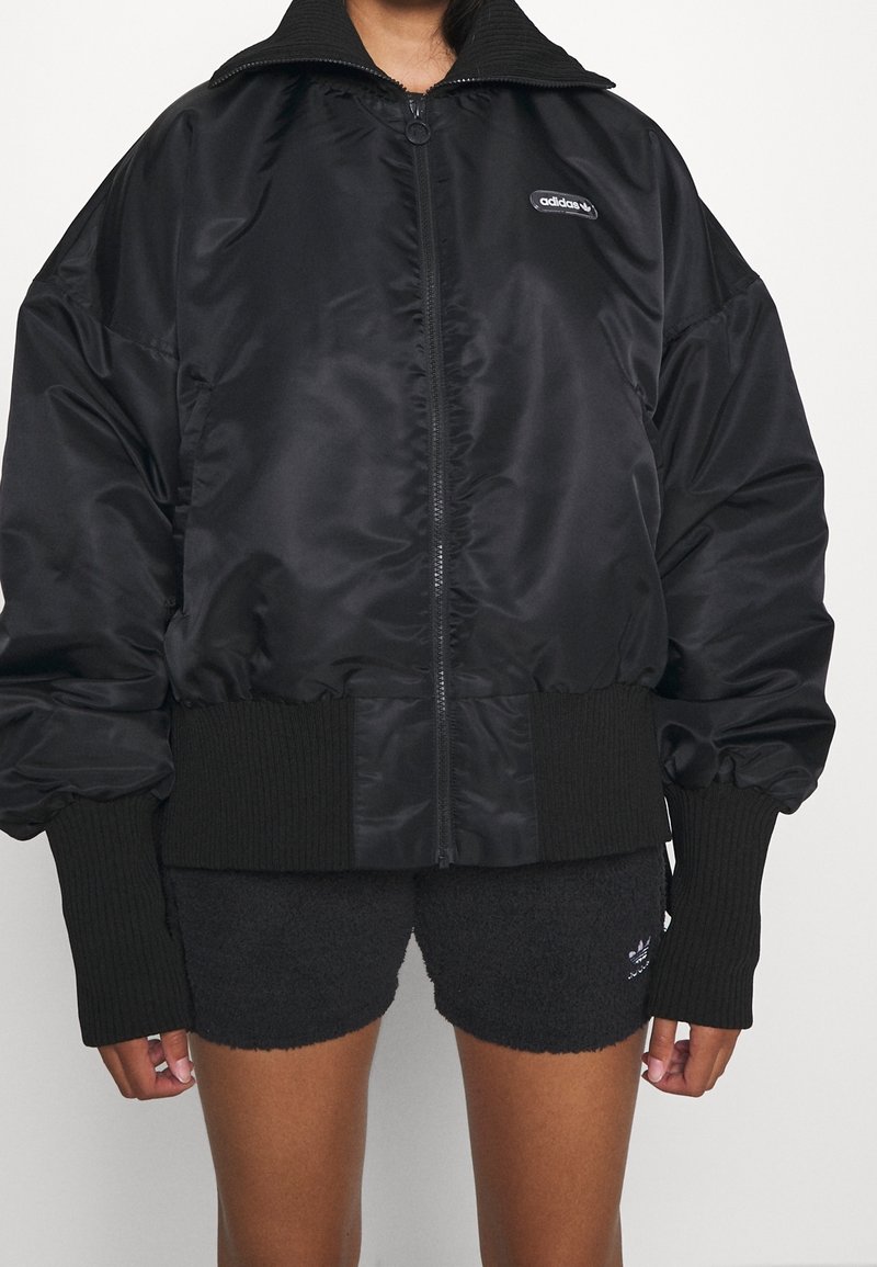 Veste noire zippée avec une finition satinée, coupe surdimensionnée, poignets et ourlet côtelés, et col montant. Présente un logo brodé sur la poitrine.