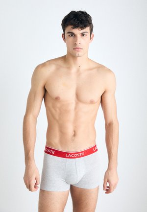 Grijze stoffen boxershorts met een rode tailleband met het Lacoste-logo. Het model is shirtloos en staat voor een neutrale achtergrond.