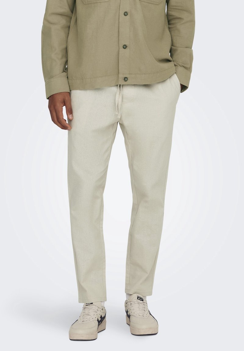 Licht beige broek met een tailleband met trekkoord, gemaakt van een textuurstof. Gecombineerd met een groene overhemd met knopen en witte sneakers.