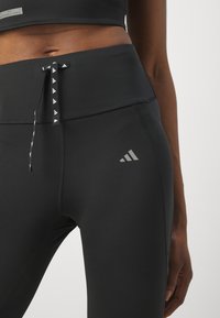 High-waisted black leggings with a smooth texture, featuring a drawstring with patterned accents and a small grey logo on the thigh.