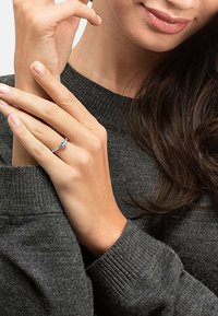 Silberring mit einem zentralen Edelstein, in einem zarten Band gefasst. Hand vor einem dunkelgrauen Pullover präsentiert, wobei die glatte Hautstruktur zur Geltung kommt.