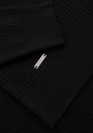 Musta teksturoitu kangas, jossa on vohvelikuvio ja ribbiedustat, jotka on täydennetty hopeisella tuotemerkkitunnuksella.