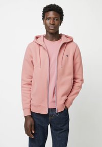 Lichtroze rits hoodie met trekkoorden, voorzien van zijzakken en een klein logo, gedragen over een lichtroze T-shirt, gecombineerd met donkere jeans.