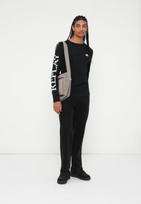 Fekete hosszúujjú póló, amelyen a "REPLAY" felirat látható az ujjon, fekete nadrág, és egy világosszürke, cipzáros részletekkel ellátott crossbody táska.