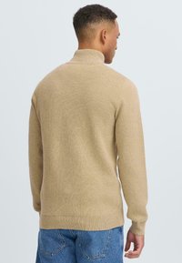 Beige gebreide trui met een hoge hals, lange mouwen, ribbels aan de onderkant en een gestructureerde stof. Standaard pasvorm, gedragen met blauwe denim jeans.