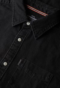 Chemise noire Superdry à boutons blancs, avec une poche poitrine unique et un détail rayé à l'intérieur du col.