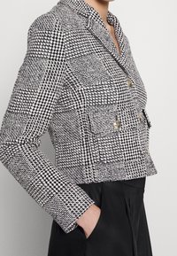 Cropped černobílá houndstooth bunda s výstřihem, dvojcestným zlatými knoflíky a dvěma předními kapsami. Texturovaná tkanina.