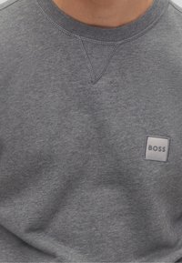 Felpa grigia in tessuto morbido con collo rotondo e dettagli di cucitura. Presenta una piccola etichetta quadrata con il logo "BOSS" sul davanti.