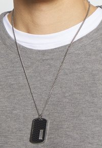 Metalowy naszyjnik z teksturowanym czarnym wisiorkiem w kształcie psa z wygrawerowanym napisem "BOSS", zawieszony na srebrnym łańcuszku, noszony na szarym swetrze.