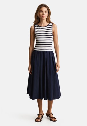 Kvinde står iført en ærmeløs kjole med en stribet hvid og navy top og en ensfarvet navy udvidet midinederdel med sorte sandaler.