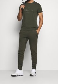 Olivově zelený sportovní outfit skládající se z trička s krátkým rukávem a úzkých tepláků, doplněný o bílé sportovní boty. Obsahuje loga značky.