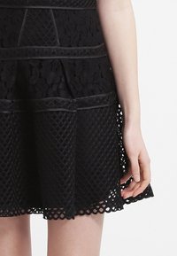 Svart klänning med texturerad spets och geometriska utskärningar. Strukturerad design med en utsvängd kjol och kontrasterande släta kantdetaljer.