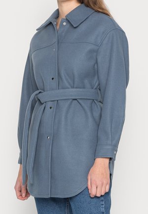 Kobieta ubrana w niebieski, wełniany płaszcz z paskiem, zatrzaskami i długimi rękawami, zestawiony z niebieskimi dżinsami.