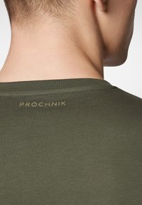 Zielona, dopasowana koszulka z gładkim materiałem. Posiada okrągły dekolt oraz nazwę marki "PROCHNIK" w złotym kolorze z tyłu.