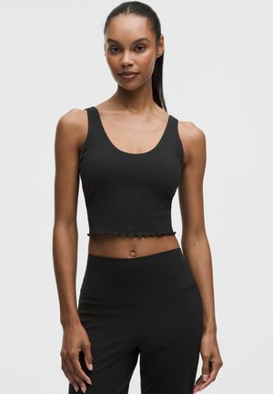 Frau mit langen Haaren, die ein schwarzes, geripptes, kurzes Tanktop und hoch taillierte Leggings trägt, vor einem schlichten hellen Hintergrund stehend.
