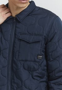 Mornarska jakna s puhanim uzorkom koja ima zatvarač sprijeda, šiljasti ovratnik, jedan džep na prsima i logotip na džepu. Glatka tekstura i lagani dizajn.