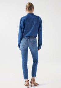 Camisa de denim en un azul profundo, de manga larga, con puños abotonados; combinada con pantalones vaqueros ajustados en un azul claro, con bolsillos traseros con detalles de costura. Zapatos de tacón color marrón.