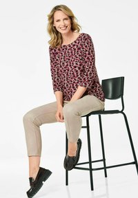 Bunt gemusterter Oberteil mit einem pinken und lila Leopardenmuster, kombiniert mit hellbeigen, schmal geschnittenen Hosen und schwarzen klobigen Loafers, sitzend auf einem schwarzen Stuhl.