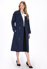 Lange marineblauwe trenchcoat met dubbelgeknoopte sluiting, grote revers en zijzakken. Glad oppervlak, knoopaccenten en opgerolde mouwen.