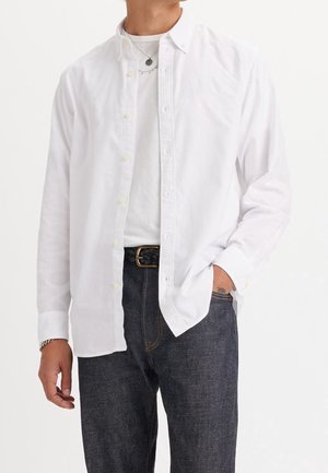 Homme portant une chemise blanche boutonnée sur un t-shirt blanc, jean denim foncé avec ceinture noire tressée, main dans la poche et bracelet argenté.