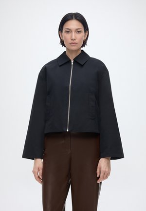 Žena s krátkymi čiernymi vlasmi, oblečená v čiernej bunde na zips a hnedých nohaviciach, stojaca pred jednoduchým svetlým pozadím.