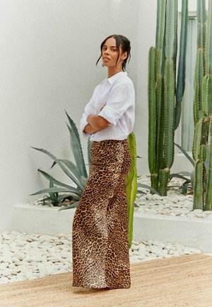 Hame leopard print yang mengalir dengan tekstur berkilau dipadu dengan kemeja putih berkancing. Latar belakang menampilkan kaktus hijau dan batu halus.