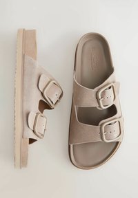 Svijetlosmeđi suede sandali s dva podesiva remena, koji imaju pravokutne kopče. Potplat je bež boje s teksturiranim uzorkom za bolje prianjanje.