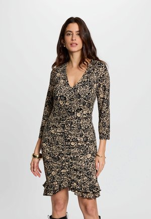Femme debout portant une robe beige ajustée avec des motifs floraux noirs, manches trois-quarts, col en V, et ourlet à volants, accessoirisée avec des bijoux en or.