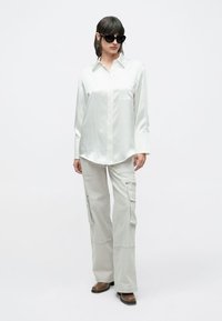 Cămașă albă de satin cu nasturi și mâneci lungi, asortată cu pantaloni cargo gri deschis cu buzunare laterale și cizme maro cu toc.