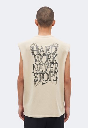 Bărbat cu păr scurt, văzut din spate, purtând un tricou bej fără mâneci cu textul „HARD WORK NEVER STOPS” și logo-ul Nike pe spate.
