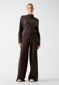 Vrouw in een donkerbruine top met lange mouwen en wijde broek, met beige schoenen, staand tegen een effen witte achtergrond.