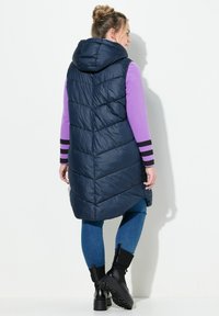 Marinblå puffväst med huva, quiltat mönster och lång längd. Parat med en lila långärmad tröja och blå jeans, svarta klumpiga stövlar.