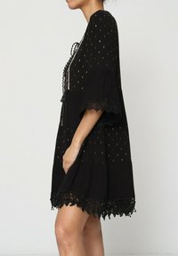 Robe noire avec des points métalliques dorés, manches courtes, avec un ourlet en dentelle et une coupe décontractée. Présente un détail à nouer au niveau du col.