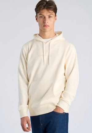 Nuori mies vaaleabeige huppari yllään ja tummansiniset farkut, seisoo yhdellä kädellä taskussa tasaisen vaaleaa taustaa vasten.