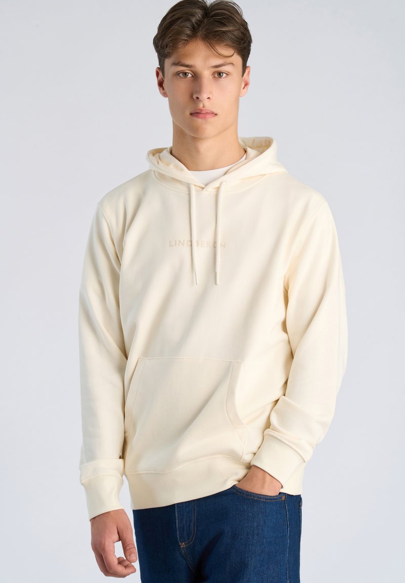 Jonge man met lichtbeige hoodie en donkerblauwe jeans, staand met één hand in de zak tegen een effen lichte achtergrond.