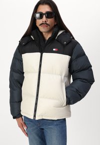 Chaqueta de plumas con capucha desmontable, sección superior negra, sección inferior crema, mangas azul oscuro, cremallera frontal y logotipo en el pecho.