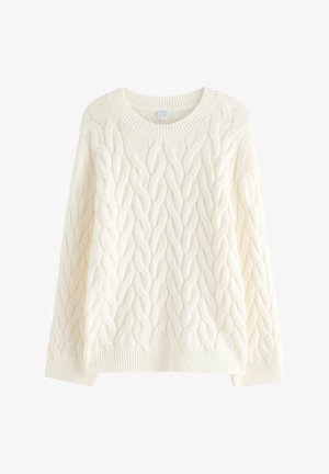 Ung person med langt krøllet hår og briller iført hvid kabelstrikket sweater, blå jeans og sorte og hvide sneakers, stående med den ene hånd i lommen.