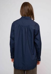 Hadseregkék hosszú ujjú ing klasszikus gallérral, hátul fodros díszítéssel és fodros szegéllyel. Sima anyagból készült.