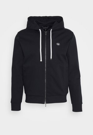 Black zip-up hoodie with a drawstring hood, featuring a logo on the chest, ribbed cuffs, and a smooth, soft fabric texture.