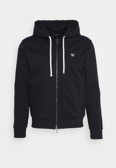 Black zip-up hoodie with a drawstring hood, featuring a logo on the chest, ribbed cuffs, and a smooth, soft fabric texture.