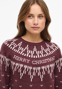 Knit sweater in burgundy with a white pattern featuring "MERRY CHRISTMAS" text, geometric designs, and a round neckline. Soft texture.