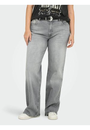 Personne portant un jean gris délavé à jambes larges, une ceinture noire cloutée, un t-shirt noir graphique et des baskets blanches, debout devant un fond uni.