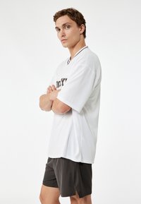 Jersey blanco de manga corta con un cuello negro y logo en la parte frontal. Lleva puestos unos pantalones cortos gris oscuro, que tienen un corte holgado y una textura suave.