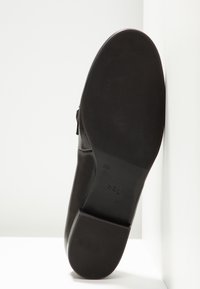 Chaussure en cuir noir à finition lisse, semelle plate texturée et petit talon. Présente le nom de la marque moulé sur la semelle.